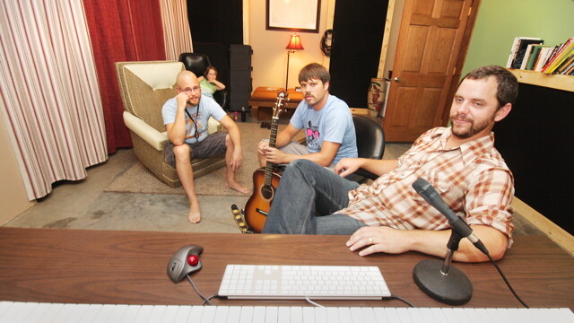 ONE KEYBOARD FOR TYPING, ONE KEYBOARD FOR MUSICING. The band Dames (Dan Boetcher, James Ignacio, and Sophie Ignacio) listen to their material recorded by Evan Middlesworth (lounging desk-side).